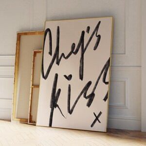 May include: A large canvas art print with the words "Chef's Kiss" in black script. The artwork is leaning against a wall, with a gold frame. A second, smaller canvas is visible behind it.