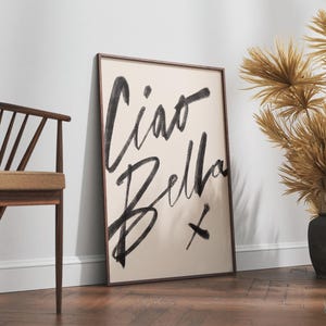 K&ouml;nnte beinhalten: Ein gerahmter Kunstdruck mit den Worten &bdquo;Ciao Bella&ldquo; in schwarzen Pinselstrichen auf cremefarbenem Hintergrund. Das Kunstwerk lehnt an einer wei&szlig;en Wand, neben einem Holzstuhl und dekorativen getrockneten Pflanzen in schwarzen Vasen.