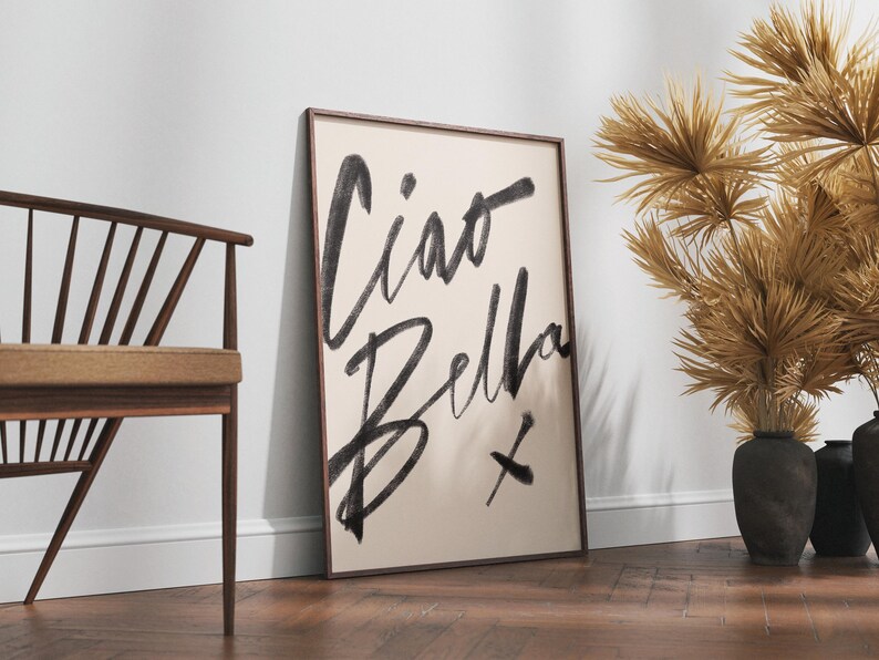 K&ouml;nnte beinhalten: Ein gerahmter Kunstdruck mit den Worten &bdquo;Ciao Bella&ldquo; in schwarzen Pinselstrichen auf cremefarbenem Hintergrund. Das Kunstwerk lehnt an einer wei&szlig;en Wand, neben einem Holzstuhl und dekorativen getrockneten Pflanzen in schwarzen Vasen.