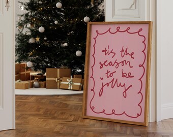 May include: A framed pink sign with the phrase "tis the season to be jolly" in red script. The sign is in a wooden frame and leans against a wall. A Christmas tree and wrapped gifts are in the background.