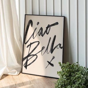 K&ouml;nnte beinhalten: Ein gerahmter Kunstdruck mit den Worten "Ciao Bella" in schwarzen Pinselstrichen auf cremefarbenem Hintergrund. Das Kunstwerk lehnt an einer wei&szlig;en Wand und einem Vorhang, mit einer kleinen gr&uuml;nen Pflanze im Vordergrund.