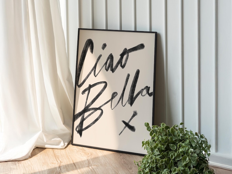 K&ouml;nnte beinhalten: Ein gerahmter Kunstdruck mit den Worten "Ciao Bella" in schwarzen Pinselstrichen auf cremefarbenem Hintergrund. Das Kunstwerk lehnt an einer wei&szlig;en Wand und einem Vorhang, mit einer kleinen gr&uuml;nen Pflanze im Vordergrund.