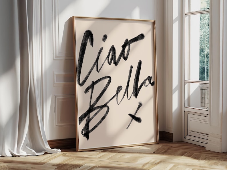 K&ouml;nnte beinhalten: Ein gerahmter Kunstdruck mit den Worten "Ciao Bella" in schwarzer Schrift auf cremefarbenem Hintergrund. Das Kunstwerk wird in einem Raum mit nat&uuml;rlichem Licht pr&auml;sentiert und lehnt an einer Wand. Der Rahmen ist goldfarben.