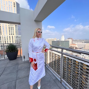 May include: A woman wearing a white dress with red trim and a floral pattern stands on a rooftop balcony overlooking a city skyline. She is wearing sunglasses and white shoes.