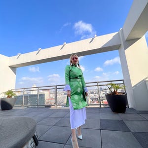 May include: A woman wearing a green and white patterned dress with a white skirt and white heels stands on a rooftop with a city view in the background.