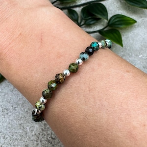 May include: A bracelet with faceted green, black, and turquoise gemstones, with small silver beads. The bracelet is worn on a wrist, highlighting the design and colour variations of the jewellery.