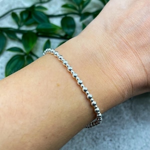 May include: A silver beaded bracelet composed of small, round beads. The bracelet is displayed on a wrist, with green leaves in the background. The beads are evenly spaced and have a polished finish.