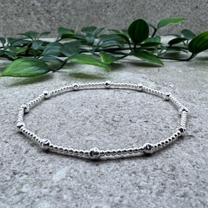 May include: A silver beaded bracelet with an elasticated design. The bracelet features small, round beads interspersed with larger, spherical beads. The jewellery is displayed on a grey surface with green foliage in the background.
