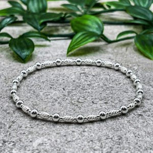 May include: A silver bracelet featuring small round beads and textured, elongated beads. The bracelet is shown on a grey surface with green foliage. The bracelet is a stretch style, suitable for various wrist sizes.