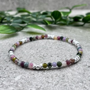 May include: A delicate bracelet with small, multi-coloured gemstone beads in pink, green, purple, and brown. The beads are strung with silver-coloured metal accents. The bracelet is displayed on a textured grey surface, with green foliage in the background.