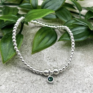 Könnte beinhalten: Ein silbernes Perlenarmband mit einem kleinen, runden, grünen Edelsteinanhänger. Das Armband besteht aus kleinen, runden, silbernen Perlen. Der Edelstein hat eine tiefgrüne Farbe und ist in eine silberne Lünette gefasst.