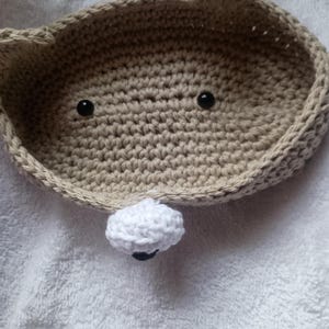 May include: A crocheted beige basket shaped like a bear with black eyes and a white crocheted nose.