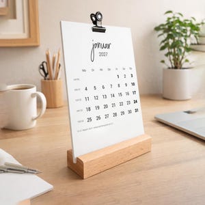 Könnte beinhalten: Ein weißer Papierkalender für Januar 2027, gehalten von einer schwarzen Metallklemme auf einem hellen Holzblockständer. Der Kalender zeigt den Monatsnamen, das Jahr, die Wochentage und die numerischen Daten in schwarzer Tinte.