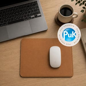 Könnte beinhalten: Ein rechteckiges Mauspad aus beigem Leder mit genähten Kanten und einer glatten Oberfläche. Eine weiße kabellose Computermaus liegt auf dem Pad. Ein silberner Laptop, eine mit dunkler Flüssigkeit gefüllte Keramiktasse und ein kreisförmiges Puk Premium-Logo sind zu sehen.