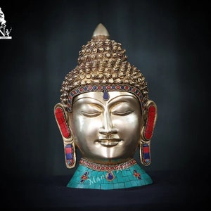 May include: A gold Buddha head sculpture with closed eyes and a serene expression. The head is adorned with turquoise and red gemstones, and the hair is styled in a traditional bun.