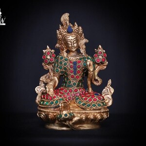 Könnte beinhalten: Eine goldfarbene Statue einer Gottheit mit einer Krone und filigranen Details. Die Statue ist mit roten und grünen Edelsteinen verziert. Die Gottheit sitzt in meditativer Haltung mit den Händen in einer Gebetsgeste.