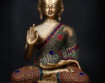 May include: A gold-coloured brass statue of Buddha sitting in a meditative pose. The statue is decorated with intricate details and colourful gemstones. The statue is set against a black background.