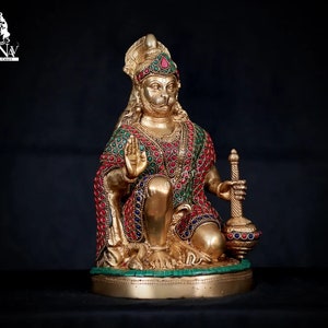 Puede incluir: Una estatua dorada del dios hindú Hanuman, sentado con la mano derecha levantada en un gesto de bendición. La estatua está adornada con piedras preciosas de colores y sostiene una maza en su mano izquierda.