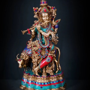 May include: A gold-toned statue of Krishna playing a flute, standing on a cow with intricate details and colorful gemstone accents. The statue is on a base with a mosaic design.