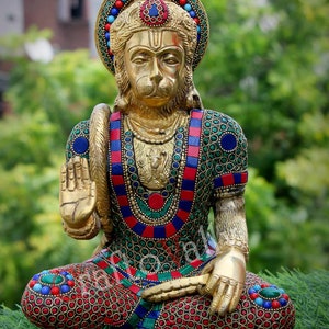 Könnte beinhalten: Eine goldfarbene Statue der hinduistischen Gottheit Hanuman, die in meditativer Pose sitzt. Die Statue ist mit bunten Edelsteinen und filigranen Details verziert. Die Statue sitzt auf einem Bett aus grünem Gras.