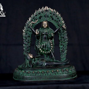May include: A green metal statue of a deity with multiple arms, standing on a figure lying on its back. The deity is surrounded by a circular frame of flames and skulls.