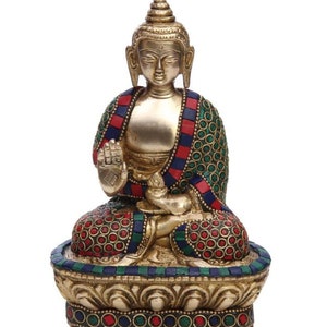 May include: A gold-colored brass statue of a seated Buddha with a red, green, and blue patterned robe. The statue is decorated with inlaid gemstones and sits on a lotus base.