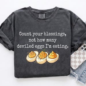 May include: A charcoal gray t-shirt featuring the white text "Count your blessings, not how many deviled eggs I'm eating." Below the text is a graphic illustration of three halved deviled eggs with yellow yolks and green garnish.