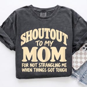 May include: A charcoal gray short-sleeve t-shirt with a crew neckline. The shirt features cream-colored text that reads SHOUTOUT TO MY MOM FOR NOT STRANGLING ME WHEN THINGS GOT TOUGH in a bold, retro-inspired font across the center of the chest.