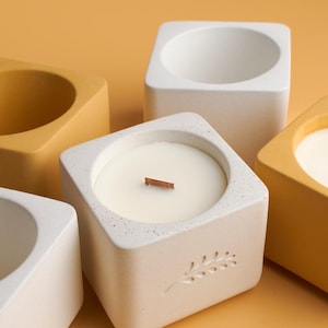 May include: Four concrete candle holders, two are white and two are yellow. One white candle holder has a wooden wick and a white candle inside. The white candle holders have a leaf design.