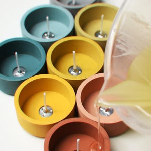 Op de afbeelding: Een set van zeven ronde betonnen kaarshouders in verschillende kleuren, waaronder grijs, blauw, geel, oranje en bruin. De houders zijn in een halve cirkel gerangschikt, en een heldere kan giet een vloeistof in een van de houders.