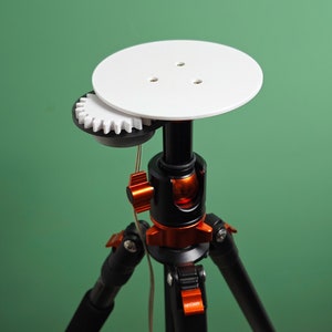 May include: A white circular platform mounted on a black tripod with orange accents. The platform has three small holes and a white gear attached to the side.