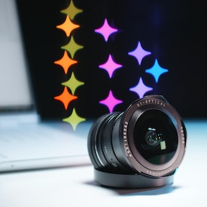 May include: A black camera lens with the text "D1-OPTICAL" and "7.5/F2.8" is positioned on a white surface. The lens is in front of a laptop and colorful star-shaped bokeh lights.