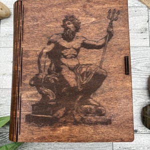 Puede incluir: Una caja de madera con tapa con bisagras. La tapa presenta una imagen grabada con láser de Poseidón, el dios griego del mar, sosteniendo un tridente. La caja está teñida de un color marrón oscuro.