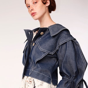 May include: A woman wearing a blue denim jacket with a white and black newspaper print skirt. The jacket has a large collar and the sleeves are gathered at the wrists.