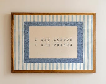 May include: A bathroom scene featuring a white bathtub, a wooden stool with toiletries, and a framed artwork with the text "I SEE LONDON I SEE FRANCE". The artwork has a blue and white striped border. The room has a neutral color palette.