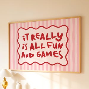 Puede incluir: Una impresión artística enmarcada con el texto "IT REALLY IS ALL FUN AND GAMES" en rojo sobre una forma blanca con bordes ondulados, sobre un fondo de rayas rosas y blancas. La obra de arte está en un marco de madera clara, colgada en una pared beige.