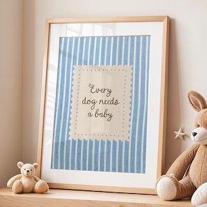 May include: A framed print with the text "Every dog needs a baby" on a blue and white striped background. The frame is light wood, and the print is displayed on a wooden table with stuffed animals.