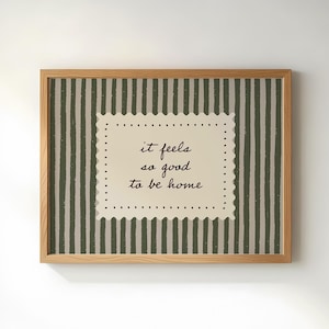 May include: A framed print with the text "it feels so good to be home." The background features vertical green and off-white stripes. The text is in a cream-coloured, scalloped-edge rectangle, and the frame is light brown.