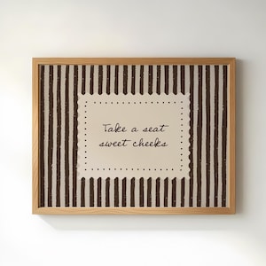Puede incluir: Una obra de arte enmarcada con un marco de madera marrón claro. La obra presenta un fondo beige con rayas verticales marrones. Un cuadrado central con el texto "Take a seat sweet cheeks" está presente.