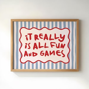 May include: A framed art print with the text "IT REALLY IS ALL FUN AND GAMES" in red, outlined in white, on a striped blue and white background. The frame is made of light-colored wood.