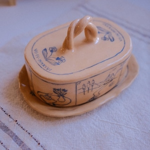 Könnte beinhalten: Beige Keramik-Butterdose mit Deckel und Unterteil, verziert mit blauen Blumen- und Landschaftsillustrationen. Der Deckel hat einen gedrehten Griff. Die Dose steht auf einer weißen Tischdecke. Maße in Zentimetern.
