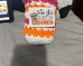 Könnte beinhalten: Ein gehäkelter Becherwärmer in Orange, Weiß und Rosa mit einem gestickten Aufnäher, auf dem "This girl runs on Dunkin'" mit einem kleinen Herz und einem stilisierten Dunkin' Donuts-Logo steht.