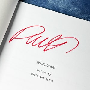 以下が含まれることがあります： 大きな赤い署名が書かれた白い脚本のページ。「THE HOLDOVERS」のテキストが中央に配置され、その下に「Written by David Hemingson」と書かれています。左上隅には、金色の「AUTHENTIC」シールとテキスト「ORIGINAL SIGNATURES, NOT A COPY / NOT A REPRINT, Certificate of Authenticity, The Holdovers (2023) Screenplay」が表示されています。