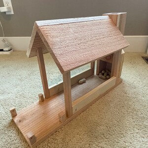 Puede incluir: Un comedero para pájaros de madera con forma de casita con techo a dos aguas. El comedero tiene una pequeña abertura en el lateral para que los pájaros puedan acceder a los cacahuetes del interior. El comedero está sobre una superficie de moqueta.