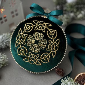 May include: A green velvet Christmas ornament with a gold embroidered Celtic knot design. The ornament is decorated with small white beads around the edge.
