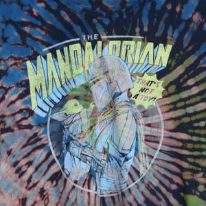 May include: Tie-dye t-shirt featuring a graphic of The Mandalorian and Grogu. The design includes the text "THE MANDALORIAN" and "THAT'S NOT A TOY!" in a circular design. The shirt has a blue, black, and orange spiral pattern.