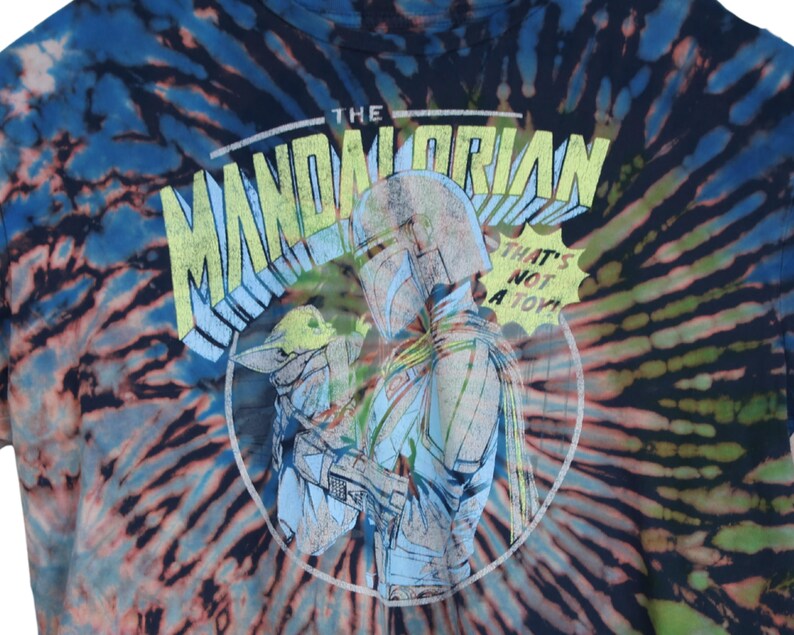 May include: Tie-dye t-shirt featuring a graphic of The Mandalorian and Grogu. The design includes the text "THE MANDALORIAN" and "THAT'S NOT A TOY!" in a circular design. The shirt has a blue, black, and orange spiral pattern.