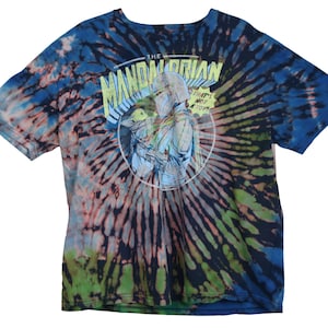 May include: Tie-dye t-shirt with a graphic from The Mandalorian. The shirt features a spiral pattern in shades of blue, green, and pink. The graphic is a circular design with the text "The Mandalorian" and "That's not a toy".