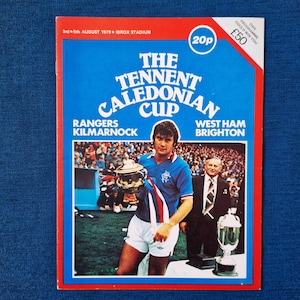 May include: A vintage football programme for the Tennent Caledonian Cup, featuring Rangers vs. Kilmarnock and West Ham vs. Brighton. The cover is in blue, red, and white, with a photo of a player holding a trophy. The price is 20p.