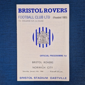 May include: A vintage Bristol Rovers football programme from 1964. The cover is white with blue text, including the team's name and details of the match against Norwich City. The programme is for the F.A. Challenge Cup.